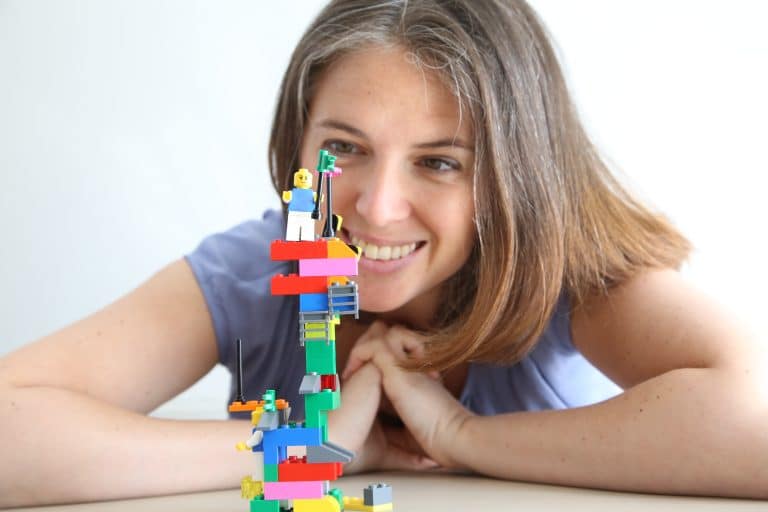 Photo of a young woman leaning against a table smiling at a LEGO Serious Play model. She is Caucasian and is wearing a blue top. The LEGO model is in focus.