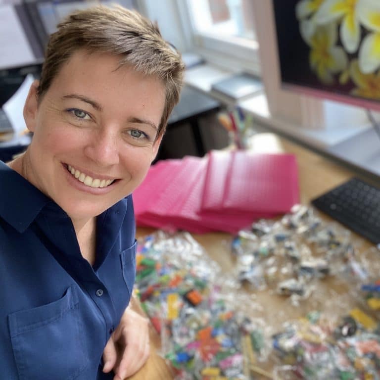 A casual photo of Joanna Maberly. She is a young Caucasian woman with short hair, wearing a blue dress. She is smiling at the camera. In the background is her work desk with pink envelopes and bags of LEGO bricks.