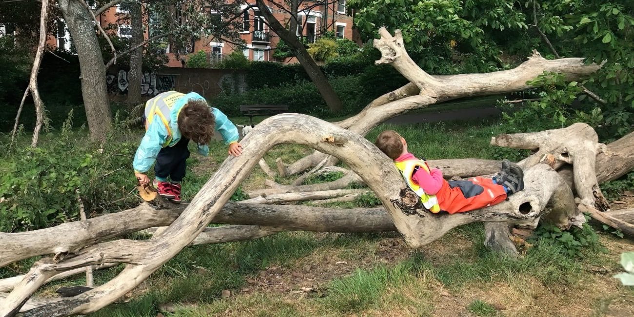 Photo of two young children playing on a large fallen tree. They are wearing brightly coloured waterproof and protective clothing and their faces are hidden. In the background is a wall with a large building behind.