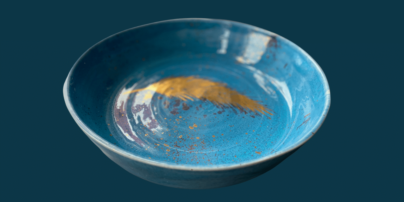 A handmade blue ceramic bowl with a golden feather painted inside. The background is plain grey-blue.