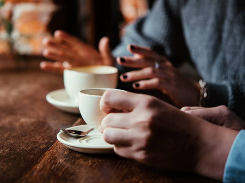 Stock image of two white women's hands while seated around a wooden table drinking coffee.