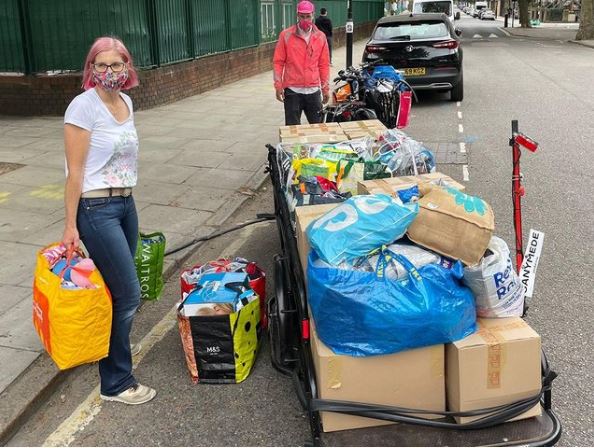 Photo at curbside showing a pedal powered courier wagon being loaded with bags and boxes. A lady in the foreground wearing a face mask and holding supermarket bags with children's items.