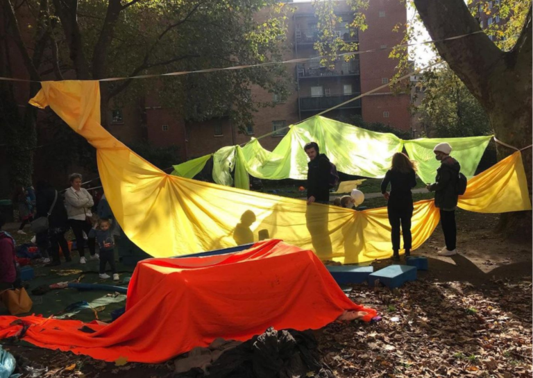 A high contrast photo of an unstructured outdoors play space. Fabrics are strung on ropes between trees and the sun is shining through the fabrics. Adults are standing around watching young children playing.
