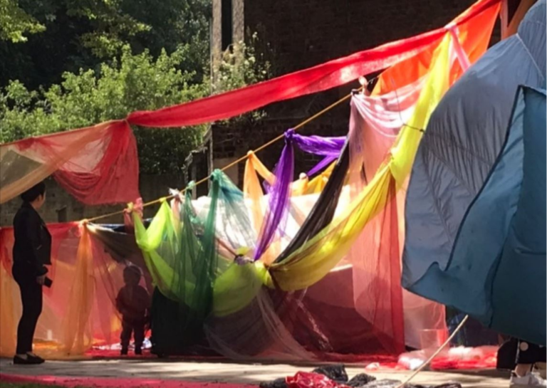 Photo of an unstructured outdoor play space. Many transparent fabrics are hung from ropes. A small child is entering the space enclosed by them. An adult watches on.