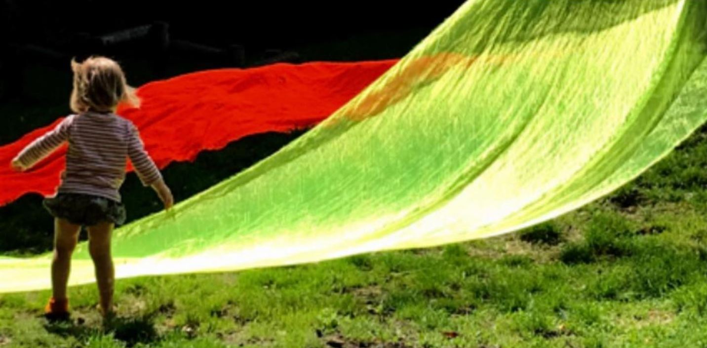 A young child stands with her back to the camera to the left of image. The rest of the image shows vivid green and red transparent play silks draped and moving in the breeze. Sunshine reflects off the green play silk.