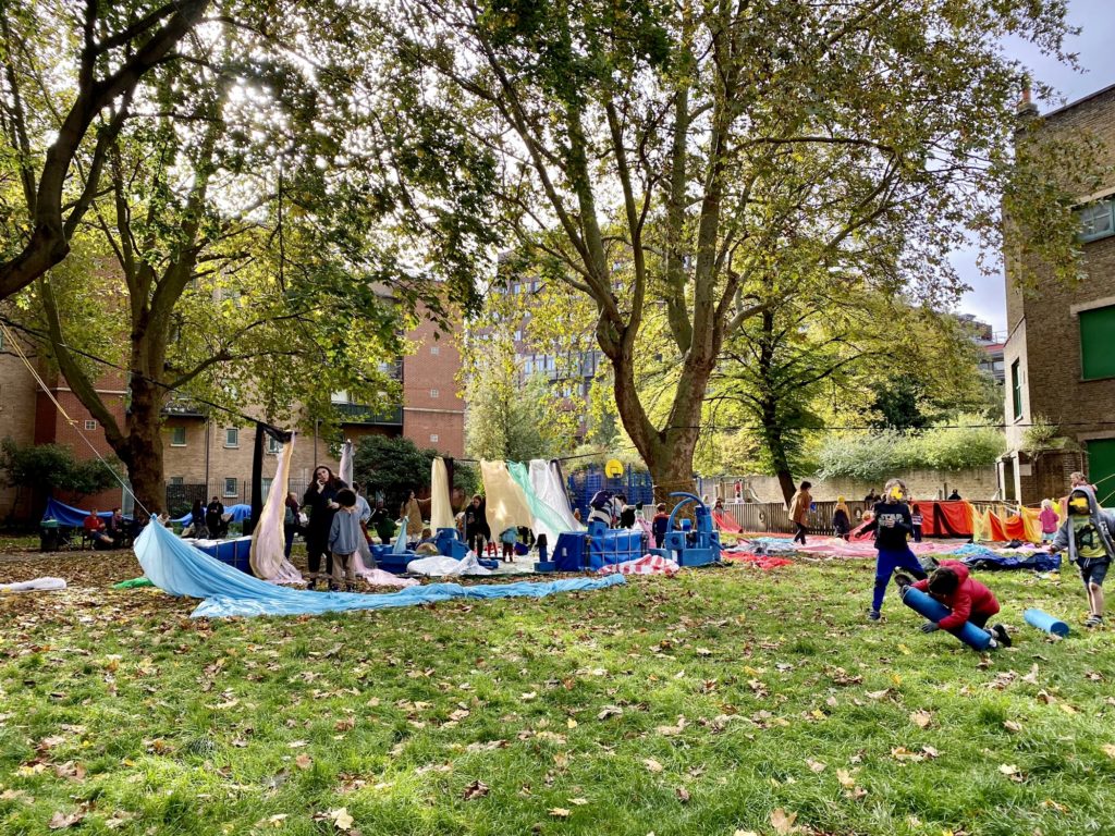 Open grassed space surrounded by residential buildings. Mature trees in the background. Children engaged in free play with loose parts, foam blocks and fabrics. The sun is shining and there are fallen leaves on the ground.
