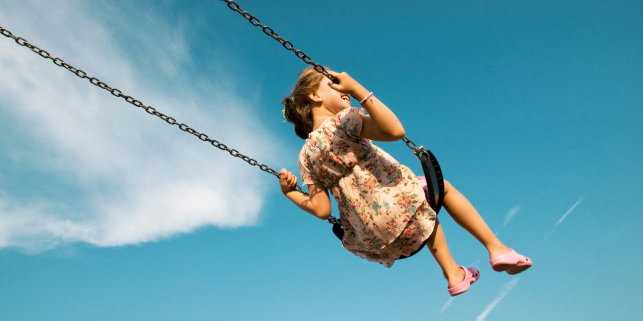 Photo taken from below of a young girl on swing with the background only blue sky and clouds.