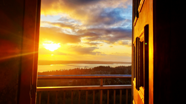 Photo of a sunset. In the foreground is an open door and a balcony railing. In the midground is the top of forest trees and a river. In the background and dominant in the image is a golden sunset sky.