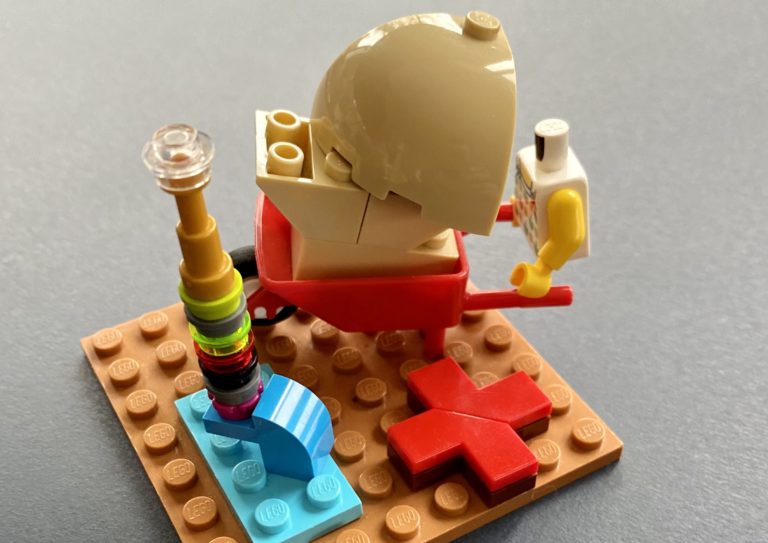 A LEGO Serious Play model shows the torso of a minifig holding a heavily laden wheelbarrow. There is a red X and a fountain on the same baseplate.
